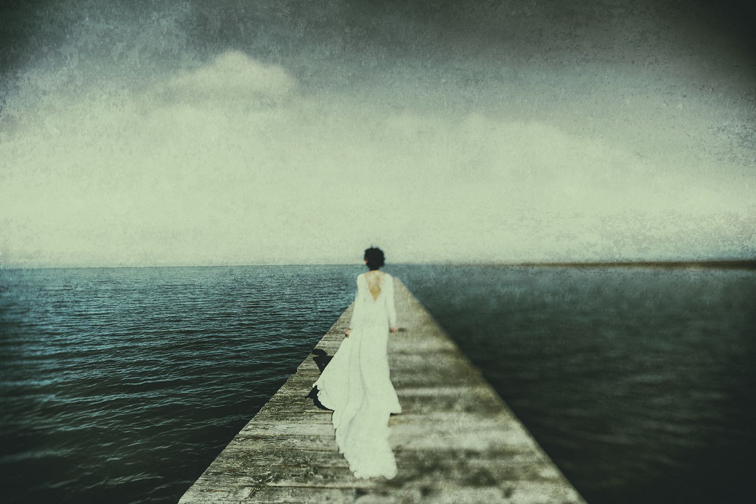 Woman in white dress standing alone on a stone pier overlooking a calm, moody ocean under overcast skies.