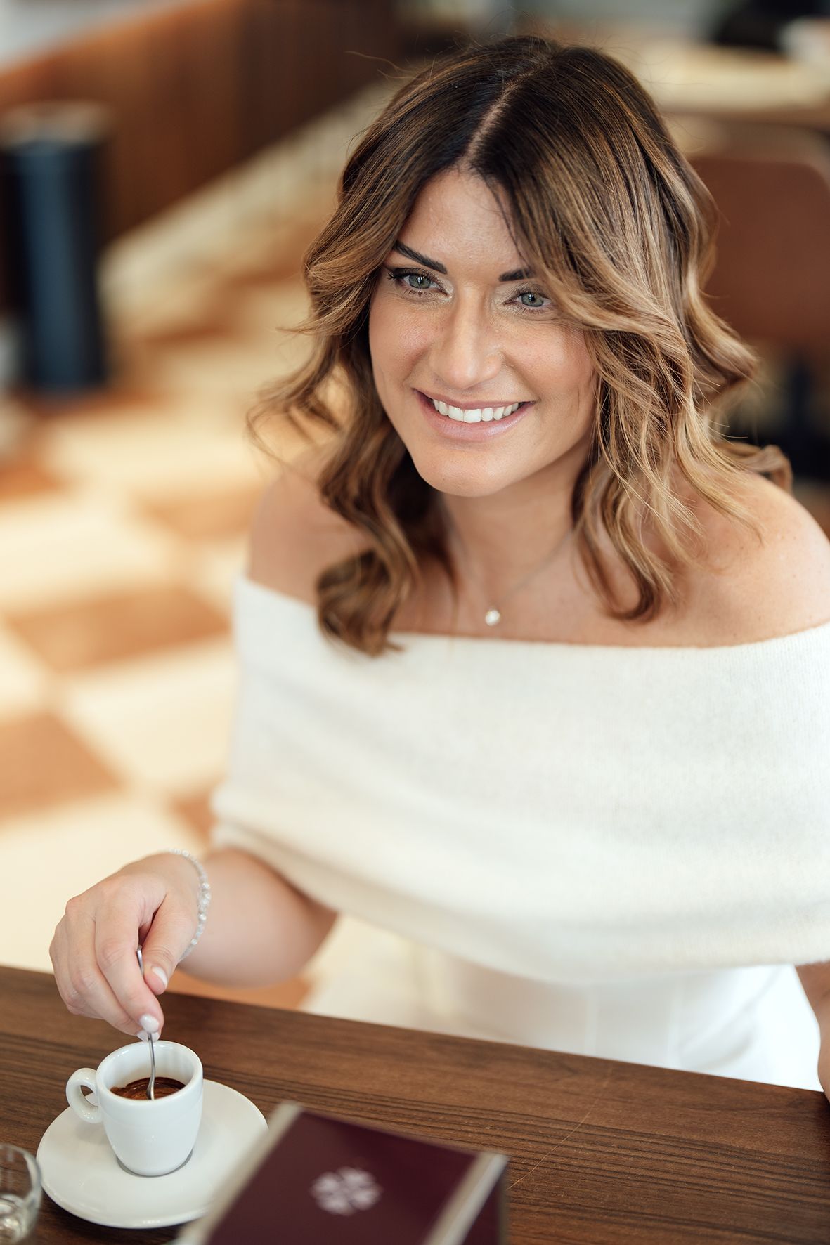 A woman in a white off-shoulder dress smiles while having coffee at a wooden table.