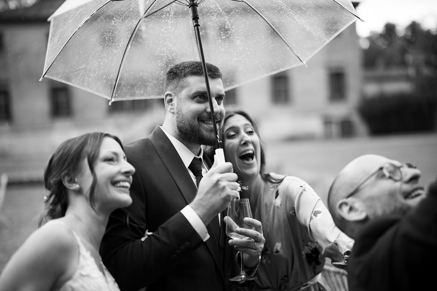 Wedding party laughing and celebrating under a white umbrella in black and white photography.