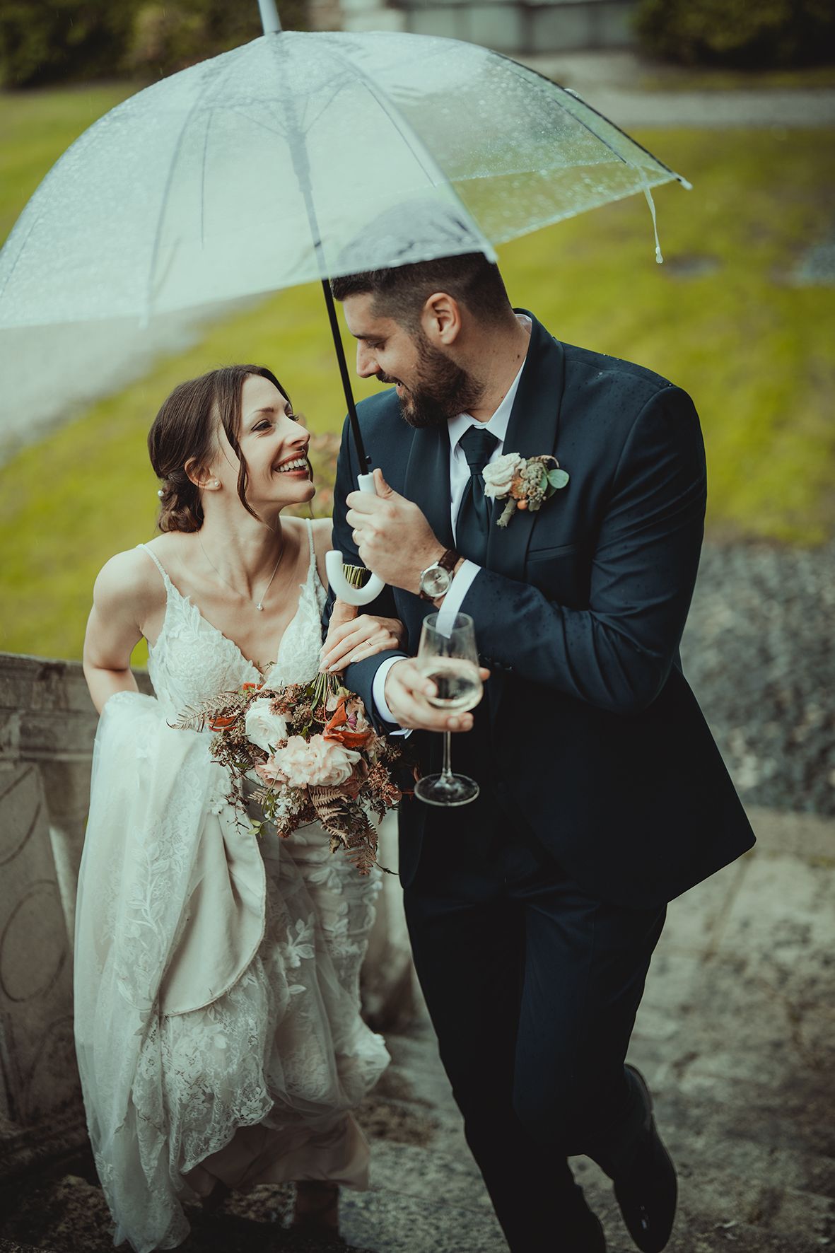 Newlyweds share champagne under white umbrellas in romantic rainy wedding photo.