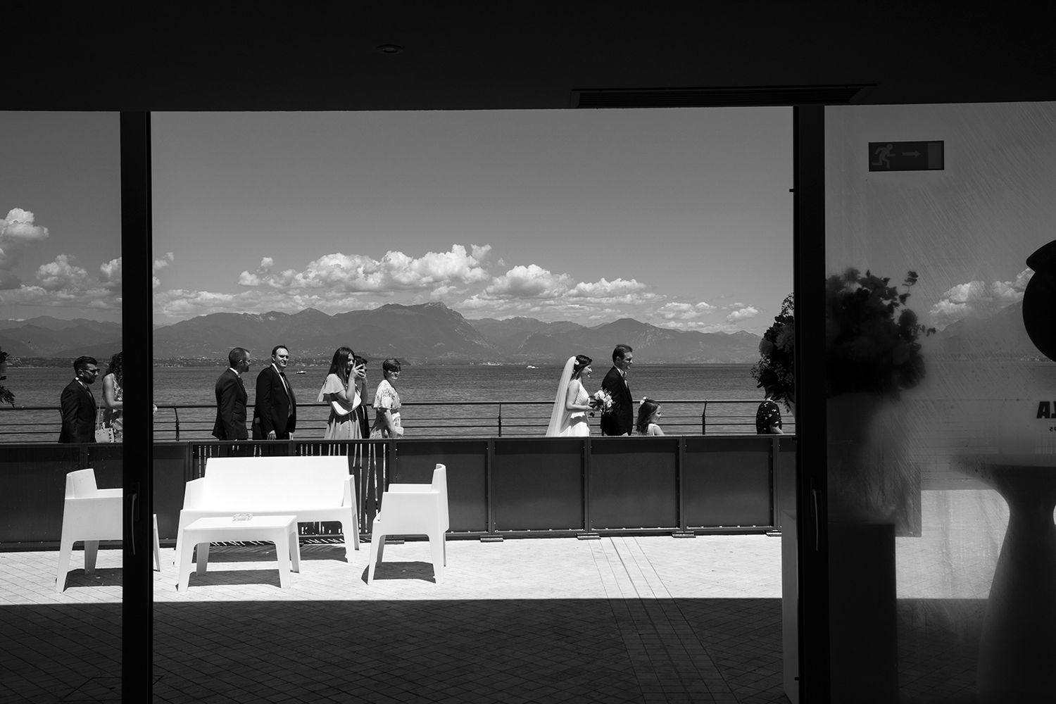 Black and white photo of elegantly dressed guests gathered on a terrace overlooking a scenic lake and mountains.