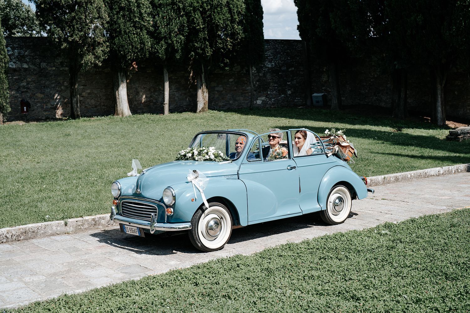 Light blue vintage convertible car parked on grass near trees on a sunny day.