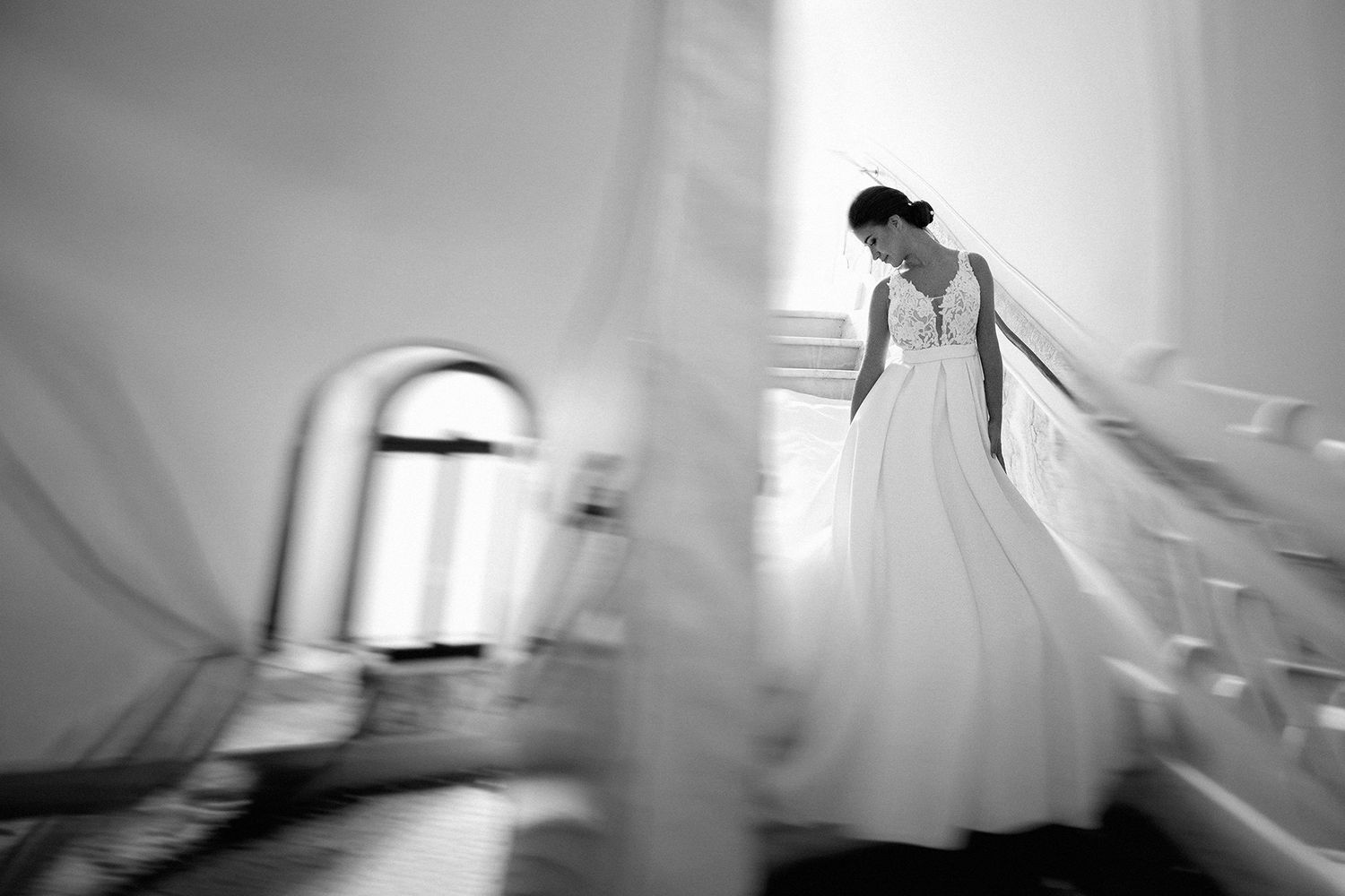 Elegant bride in a flowing white wedding gown descending a grand staircase in a black and white photo.