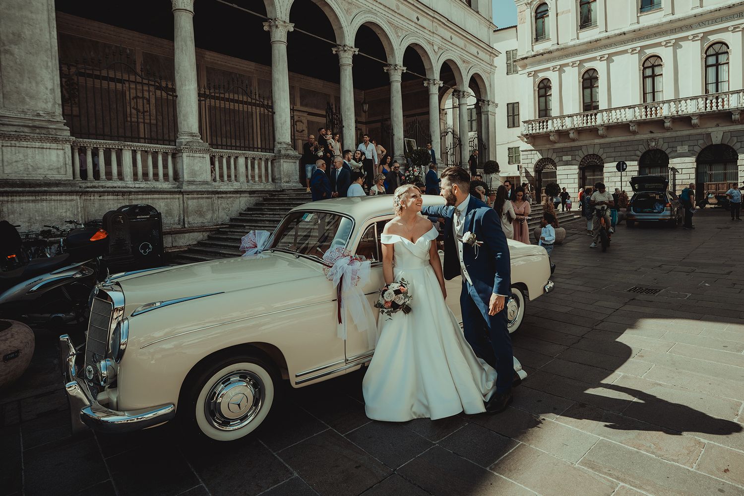 Classic car and wedding party outside a historic building with elegant architecture.