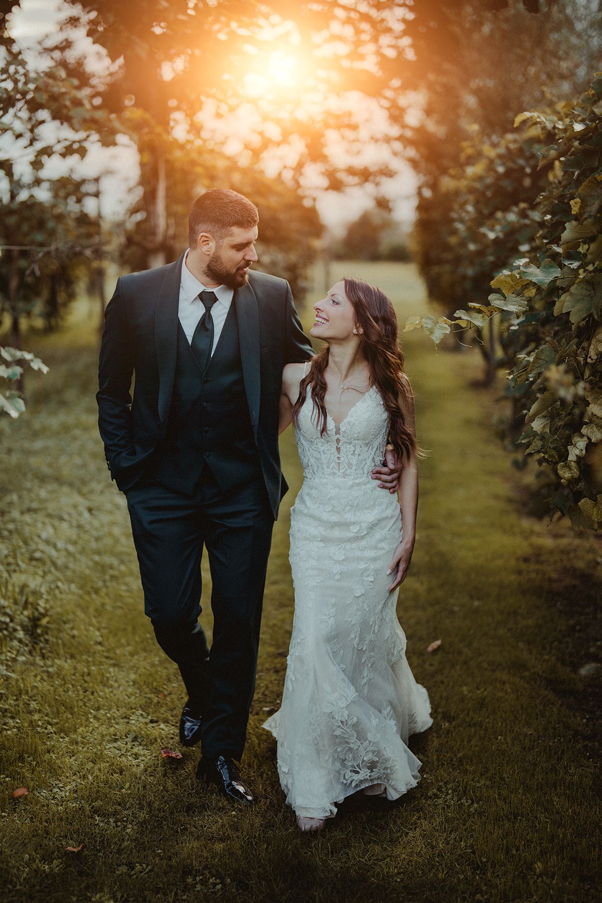 A couple walks through a sunlit vineyard at golden hour in formal wedding attire.