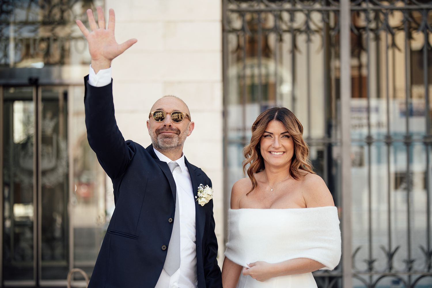 A happy couple in wedding attire poses outside, with one person waving to the camera.