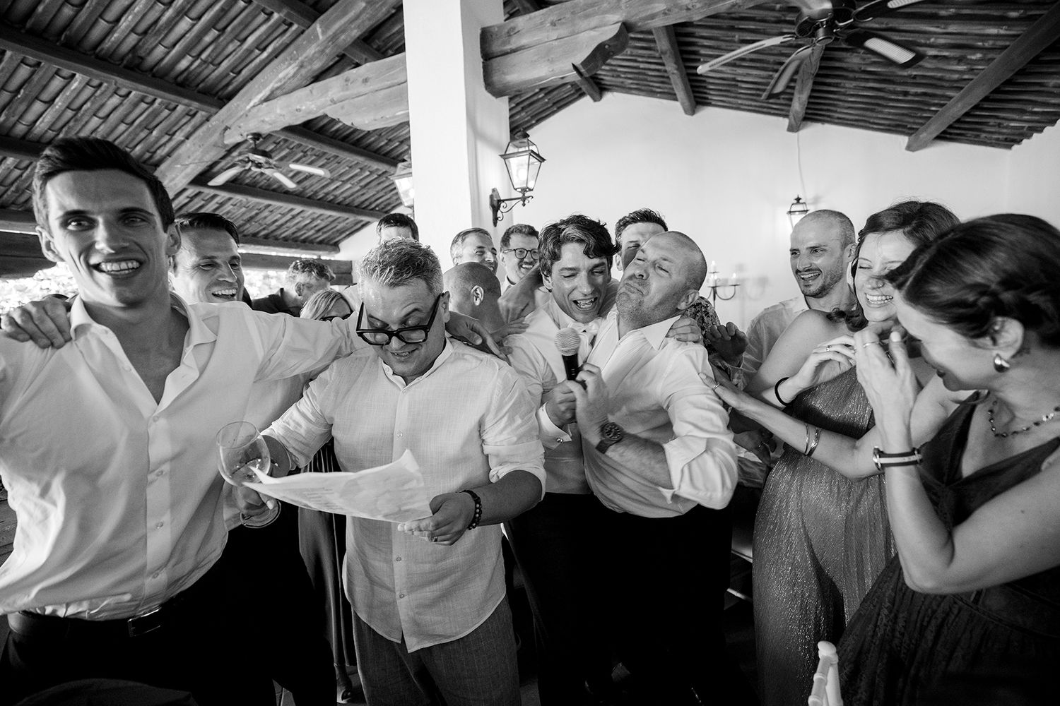 Wedding party members dance and pose under a rustic wooden ceiling at a reception.