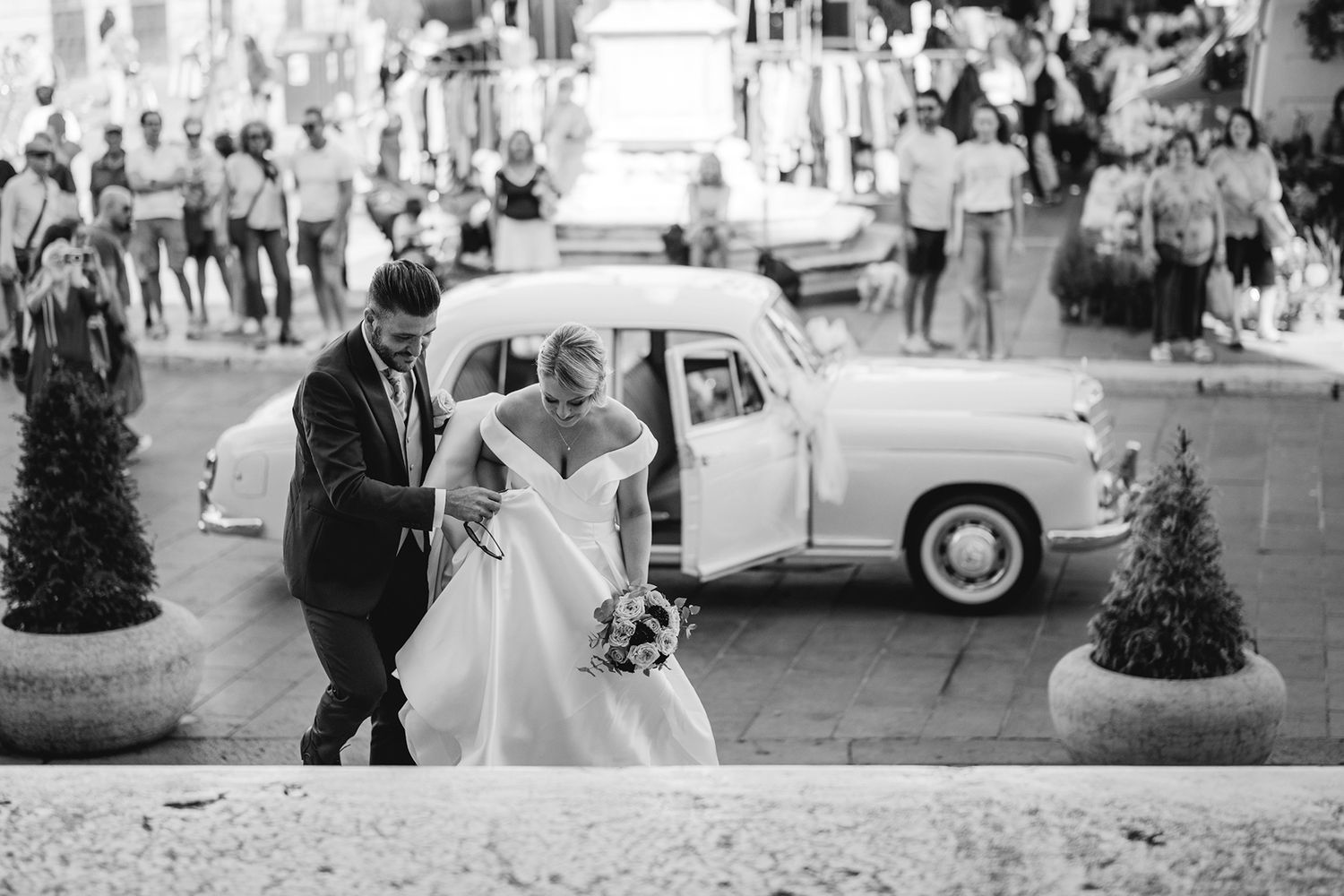 Black and white wedding photography showing guests and a vintage car outside.