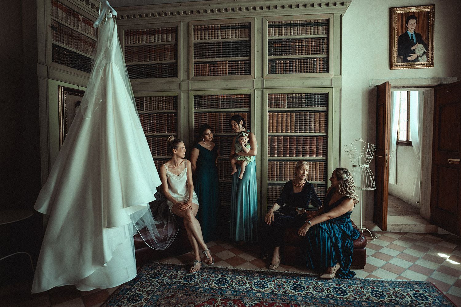Bridal party gathered in an elegant library room with a wedding dress hanging nearby, surrounded by bookshelves.