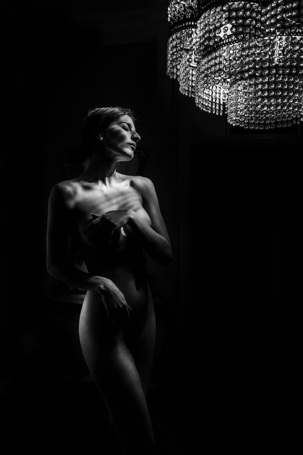 Black and white fine art nude portrait of a woman posed elegantly beneath a crystal chandelier in dramatic low-key lighting.