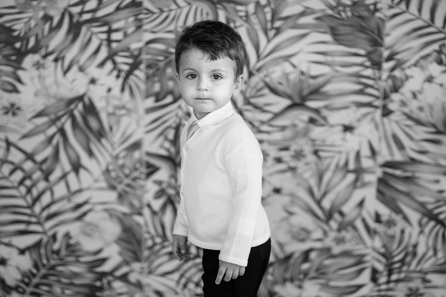 Child sitting against tropical leaf patterned wallpaper background.