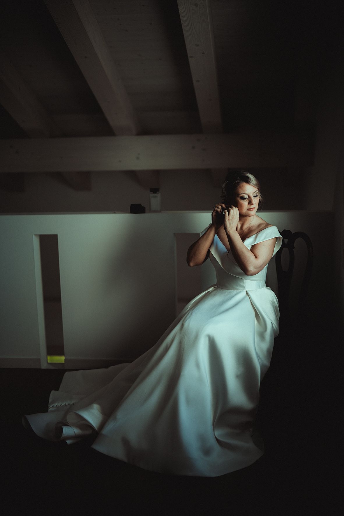 Bride in elegant white gown sits in moody lighting under wooden beams.