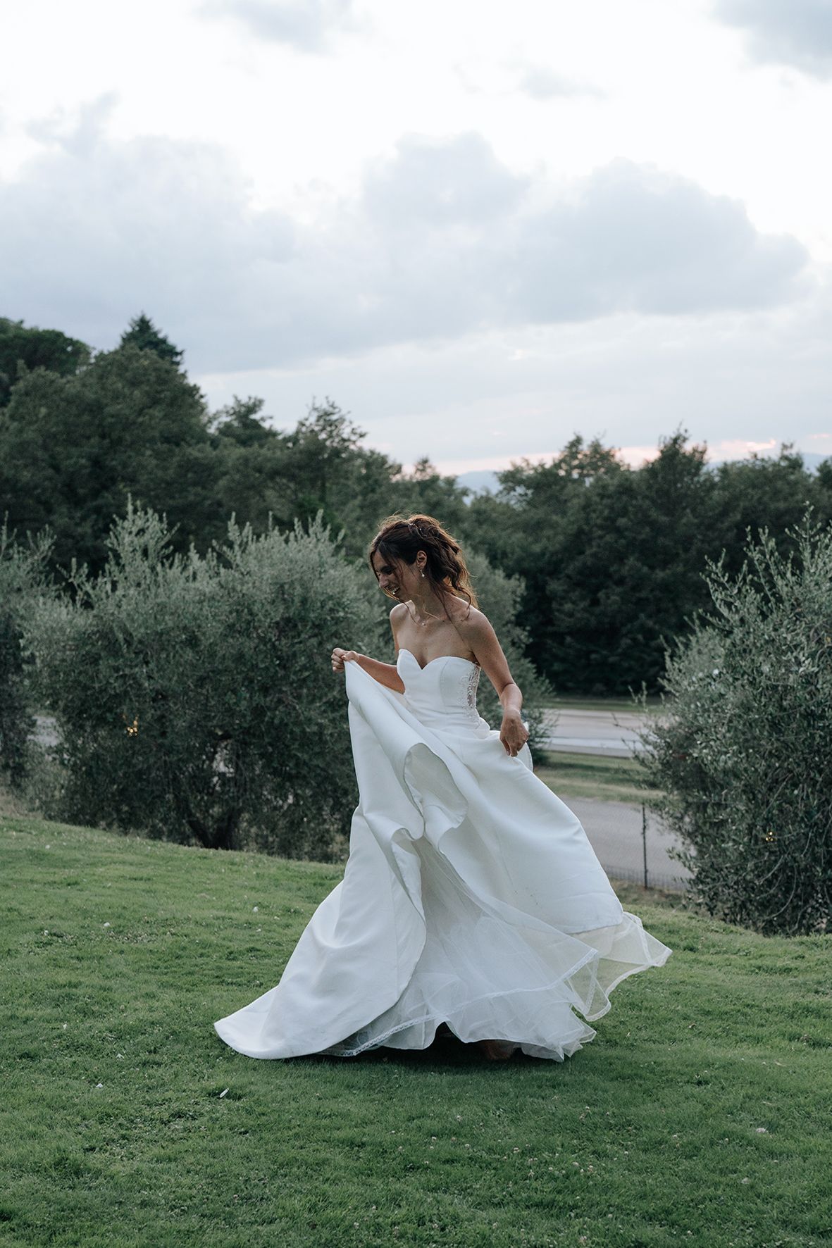 Bride in white gown dancing gracefully on a grassy hillside surrounded by olive trees.