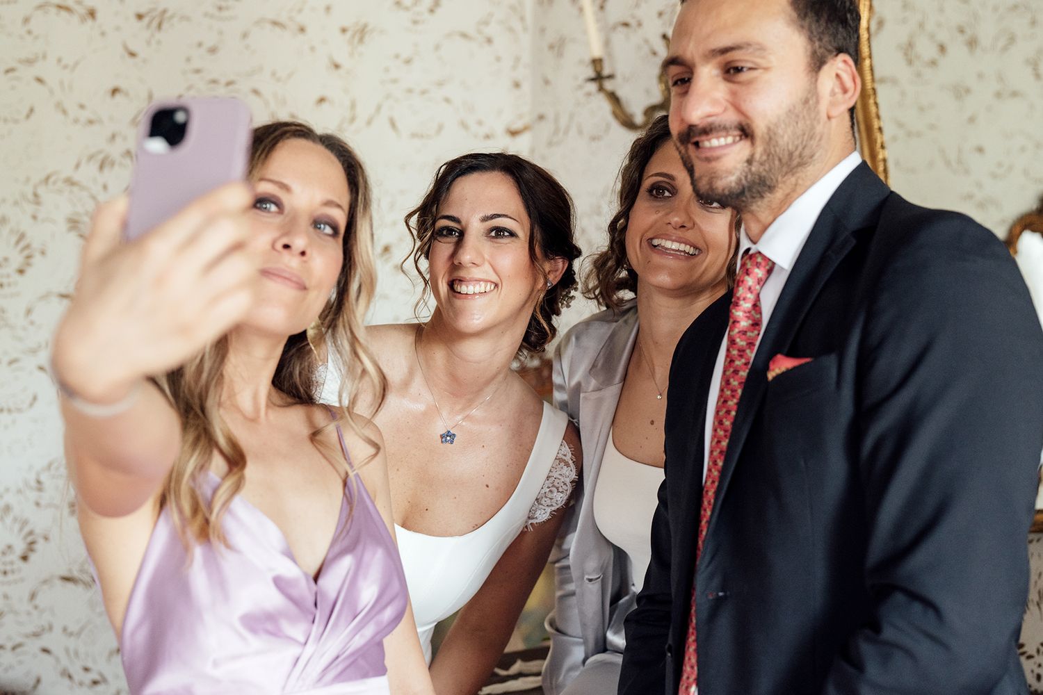 Wedding party members sharing joyful moments in a light-filled room with vintage wallpaper.