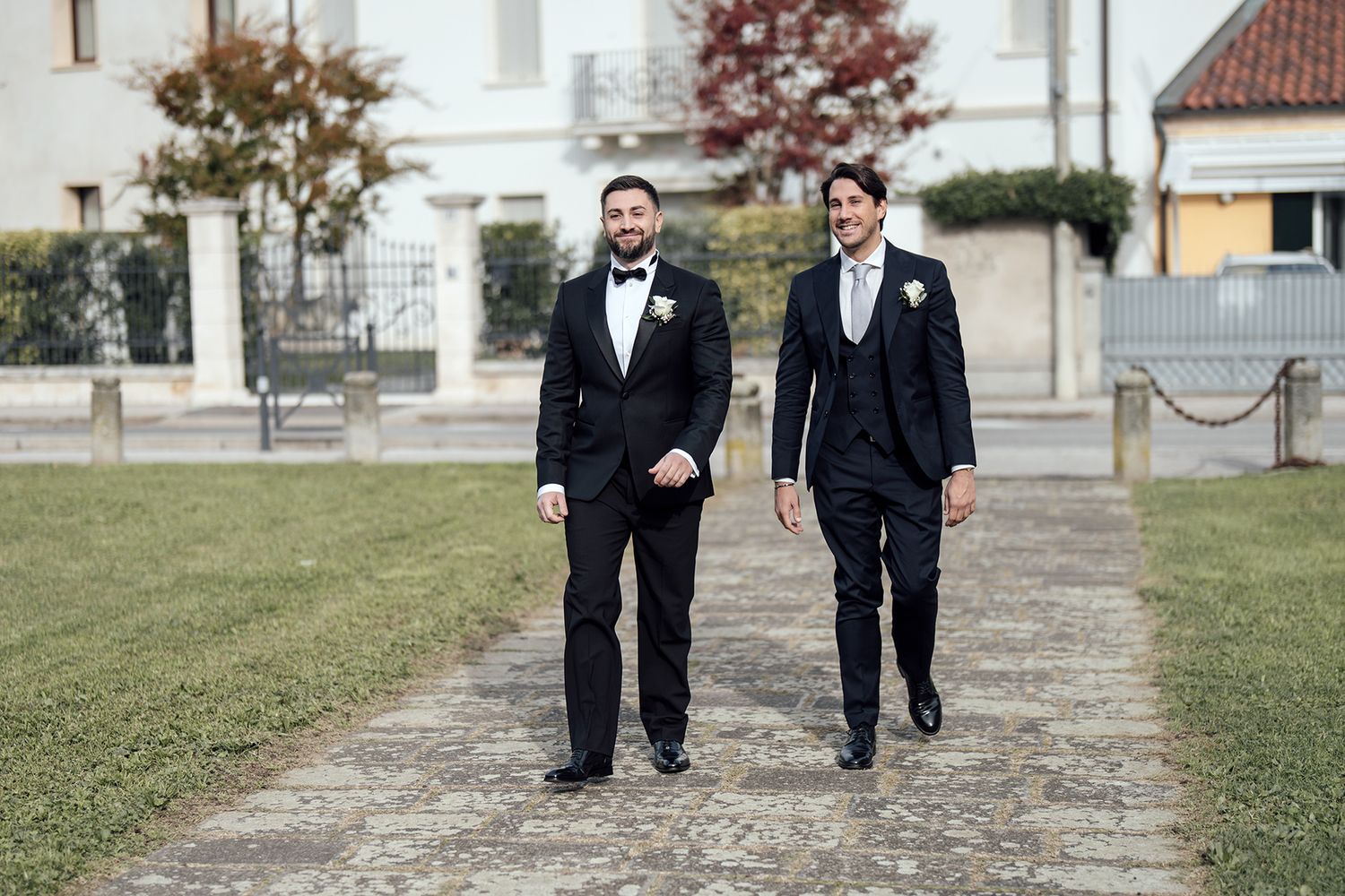 Two grooms in classic black tuxedos walk together on stone path outside church.