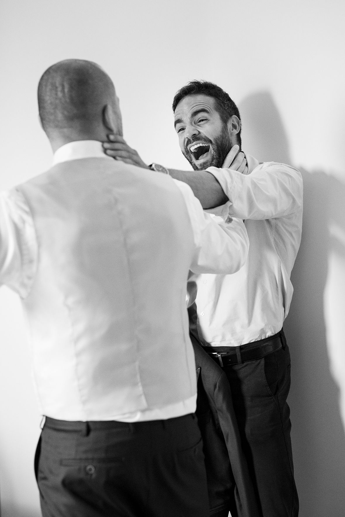 Two men in formal attire share a joyful moment while getting dressed.