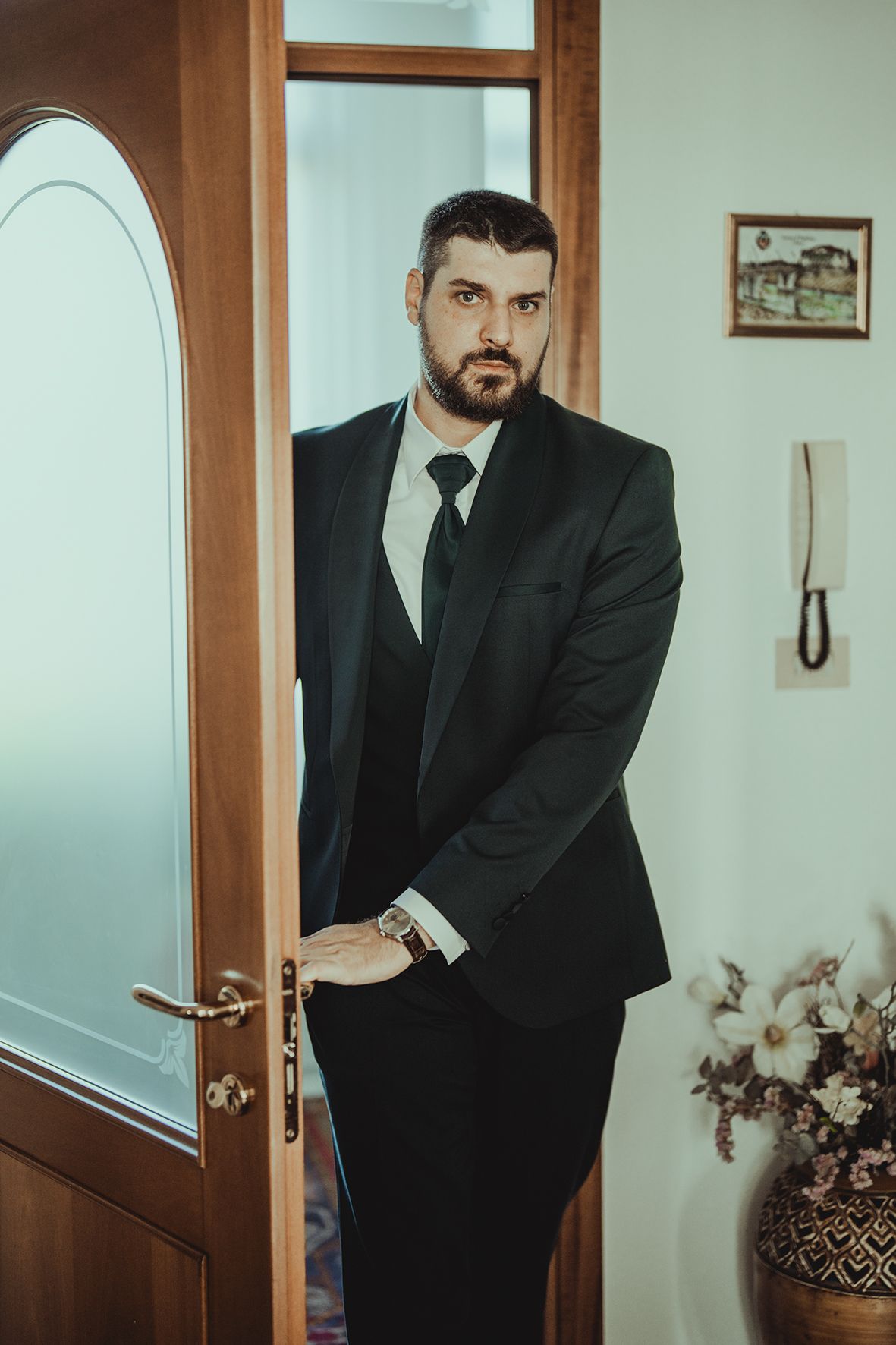A person in a black suit poses against a white wall with an arched doorway.