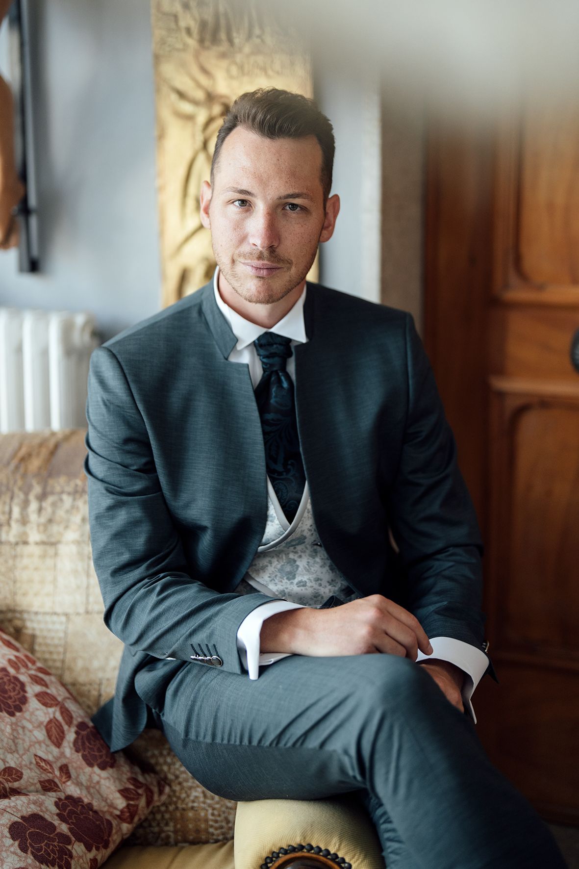 Groom in dark suit seated on vintage furniture in elegant indoor setting.