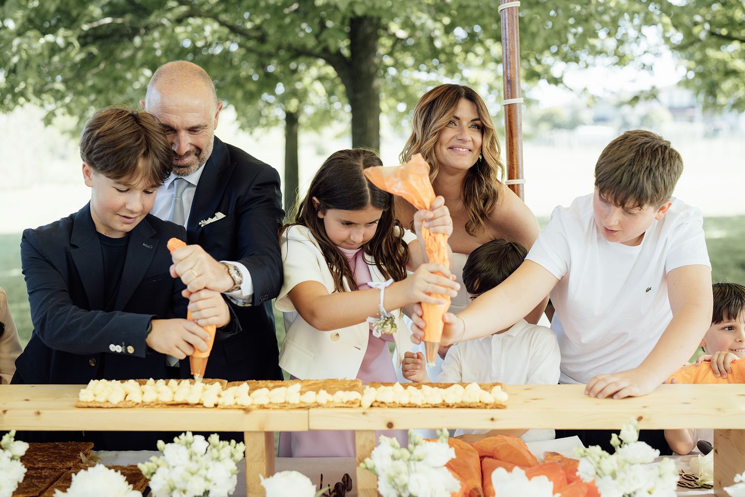 Wedding guests gathered around a table sharing desserts and laughter outdoors.