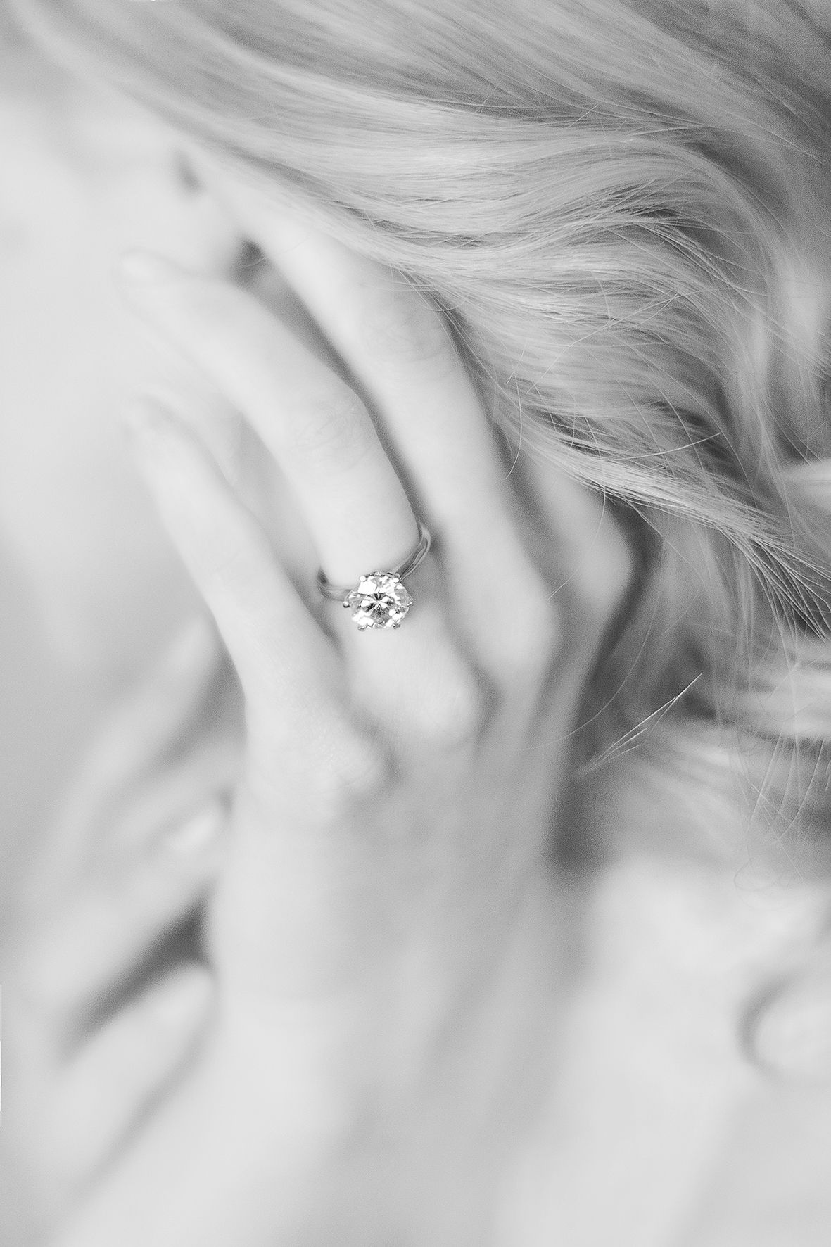 Close-up black and white photo of an elegant diamond engagement ring on a hand with soft blonde hair in background.