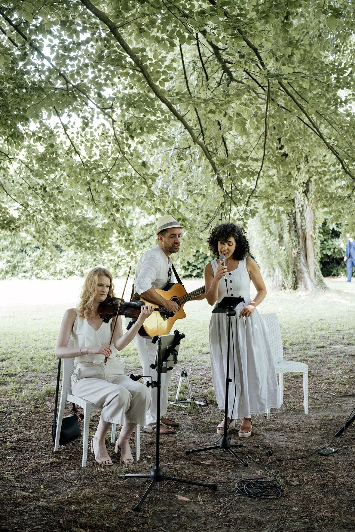 Musicians in white attire perform under trees at outdoor wedding ceremony.