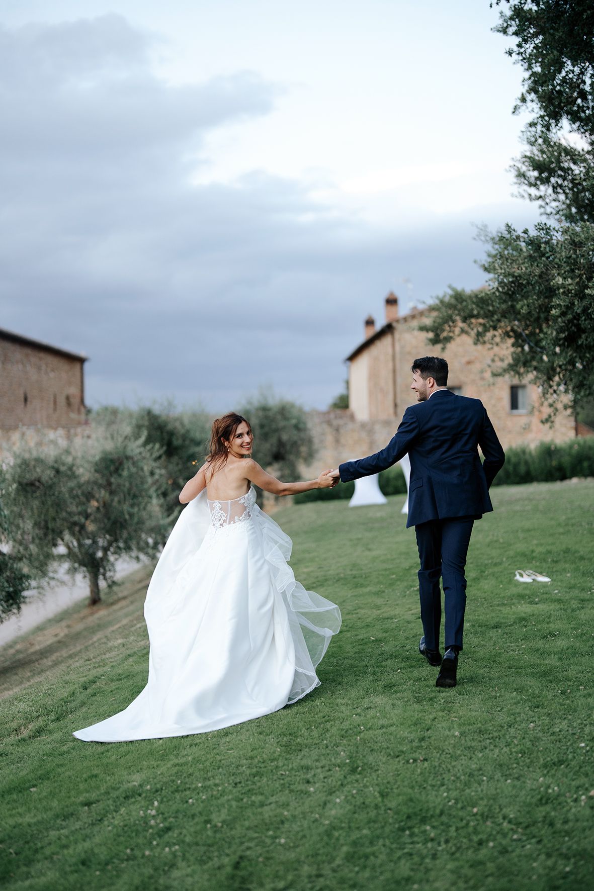 Couple walking hand in hand on a grassy hill with rustic Italian buildings in the background.