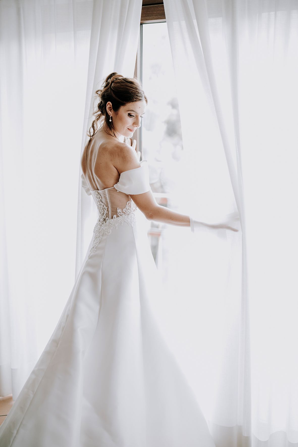 Bride in flowing white gown poses gracefully against sheer curtains in natural light.