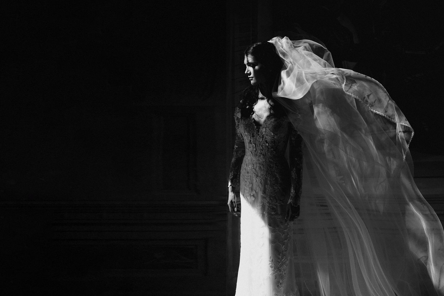 Black and white photo of a bride in a lace wedding gown with a flowing veil caught in the wind, dramatically lit against a dark background.