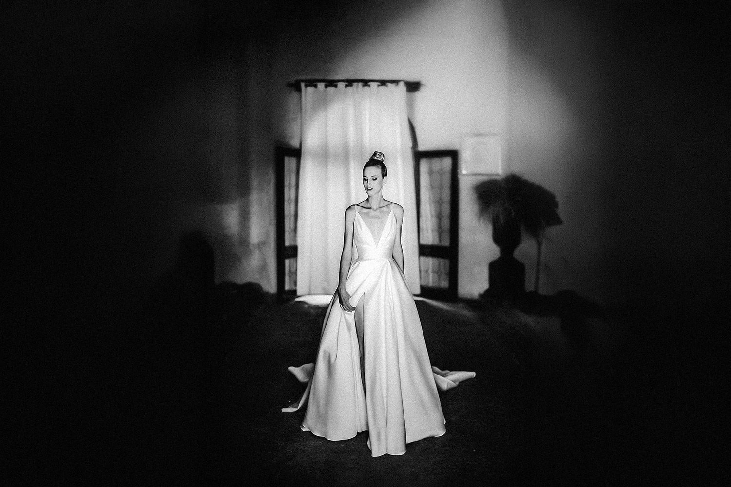 Elegant bride in a long white gown stands gracefully in a dramatic black and white portrait with soft window light.