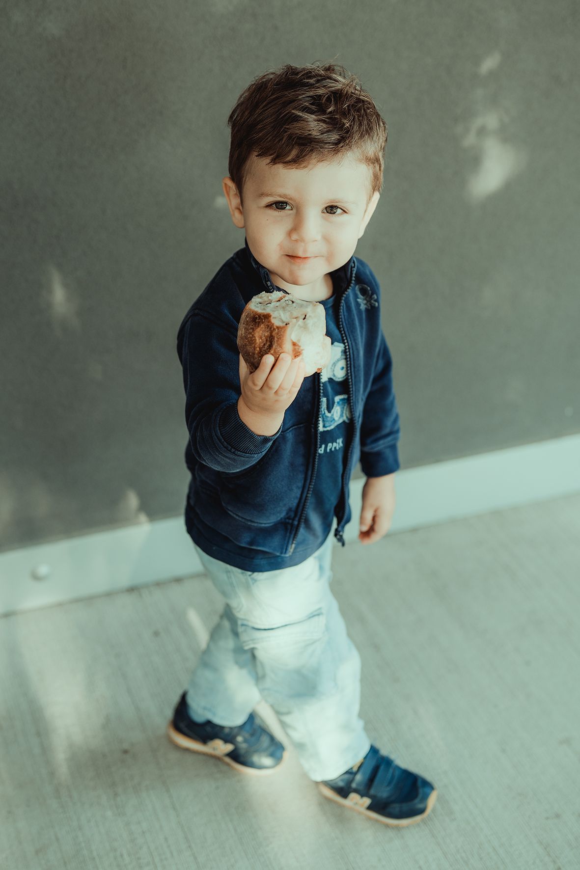 Small child in navy jacket and white pants eating a snack against gray wall.