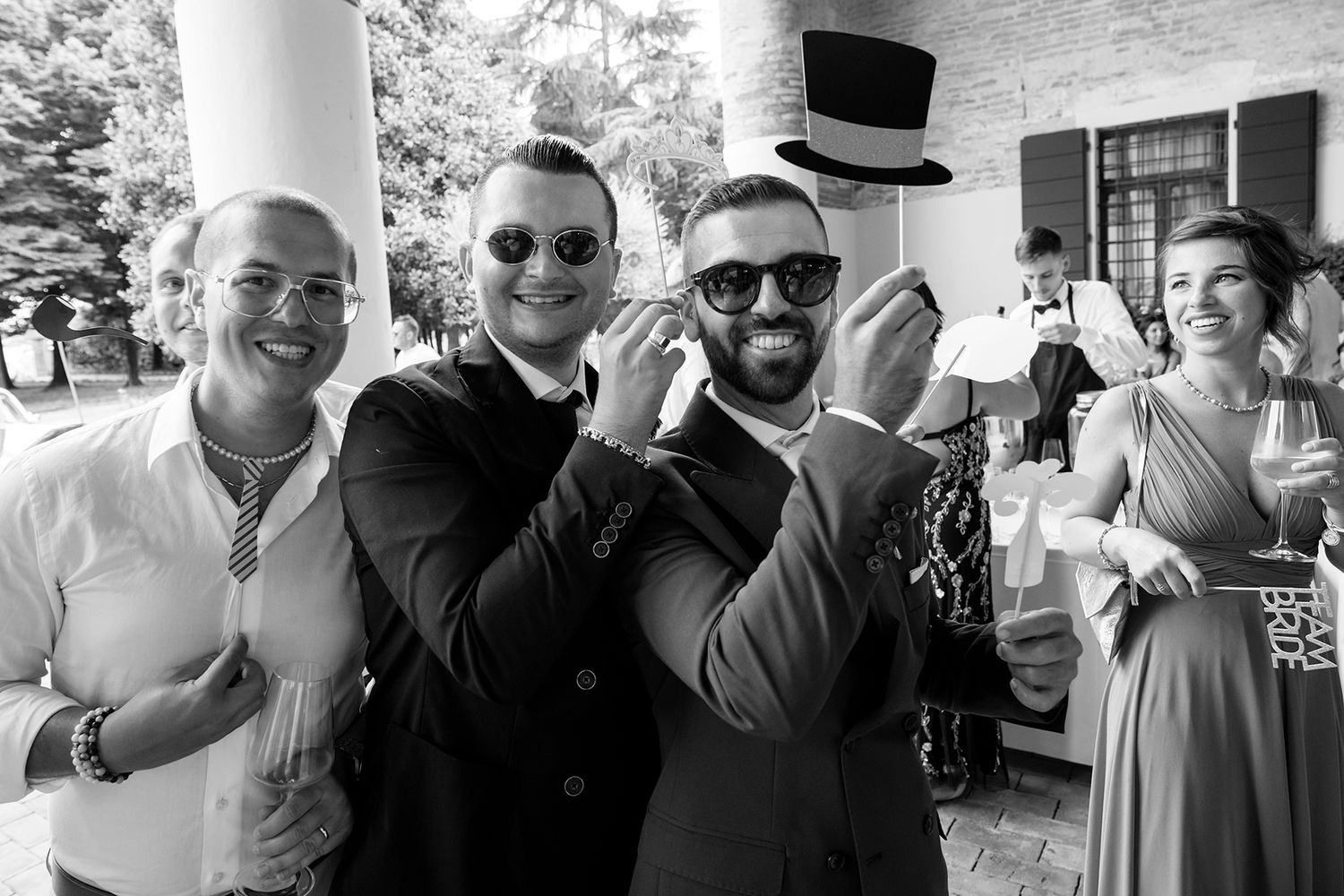 Wedding guests in formal attire sharing laughs and drinks during outdoor reception celebration.