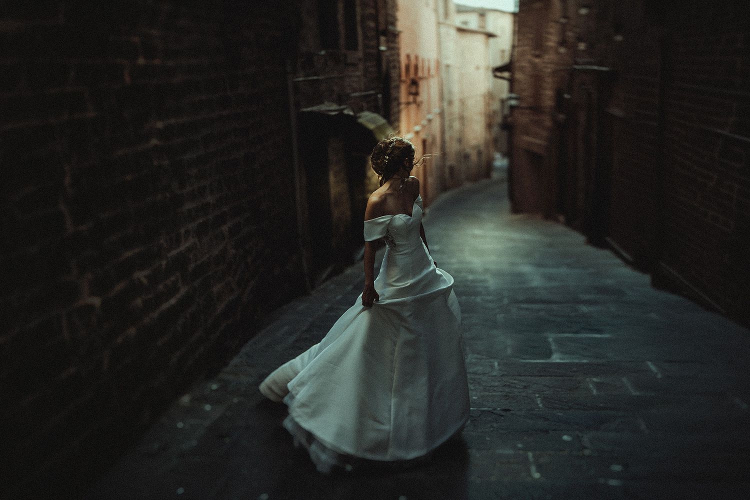 Bride in white off-shoulder ball gown walking through a narrow cobblestone alley between old brick walls.