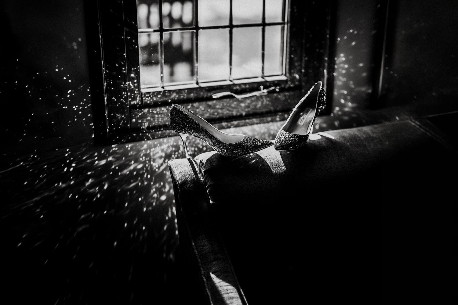 Black and white photo of legs resting on a sofa near a dusty window with light particles floating in the air.