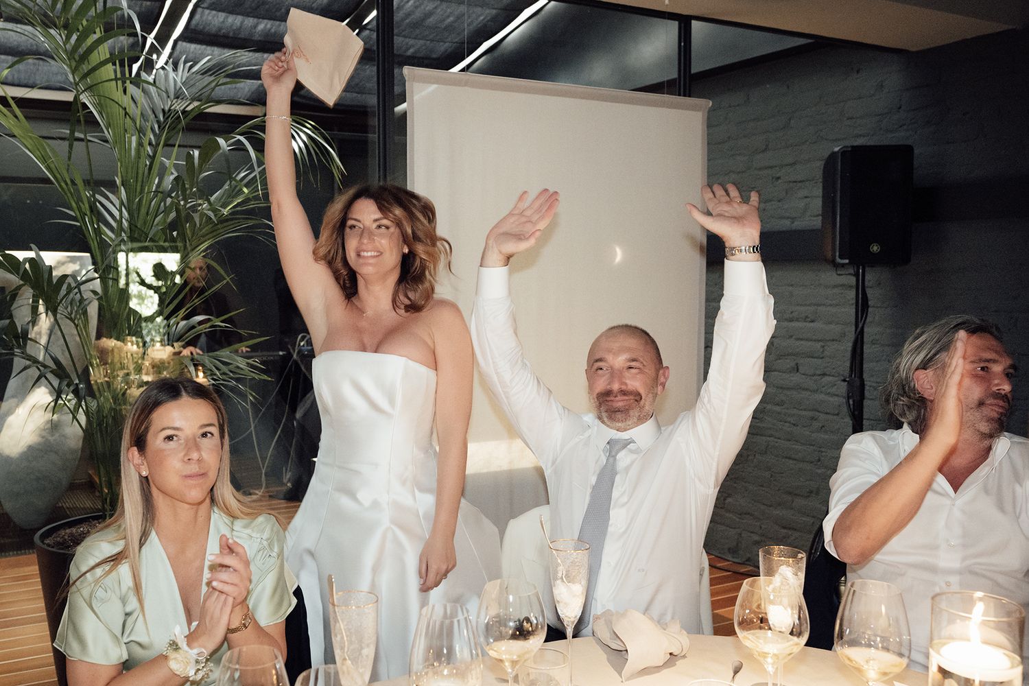 Wedding party celebrating joyfully with raised hands and drinks at an indoor reception.