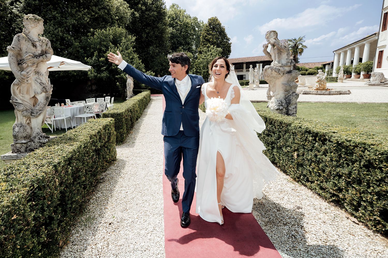 A couple walks along a gravel path surrounded by manicured hedges and stone statues in an elegant garden setting.