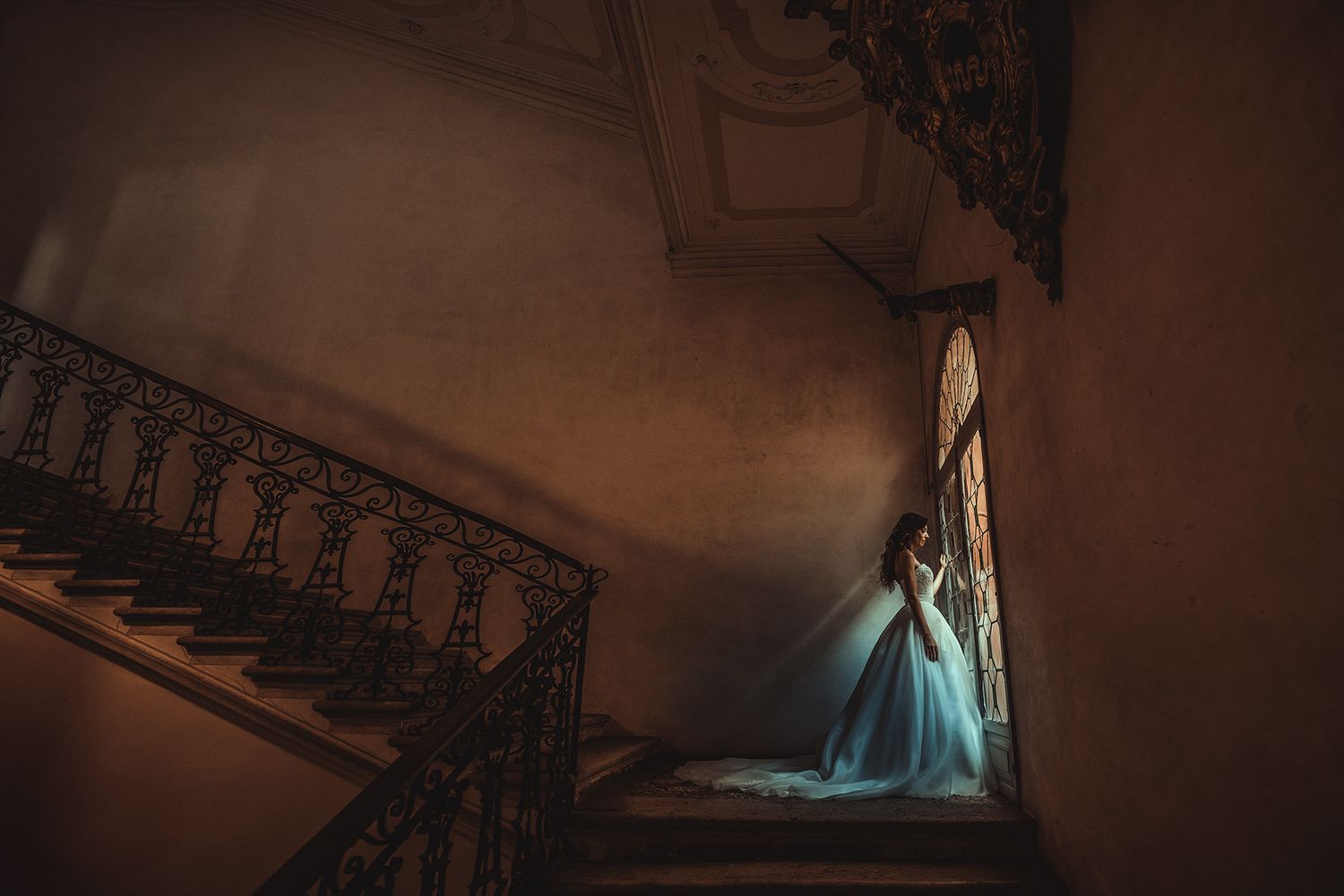 Bride in a flowing blue gown stands on an ornate staircase in a dimly lit historic mansion.
