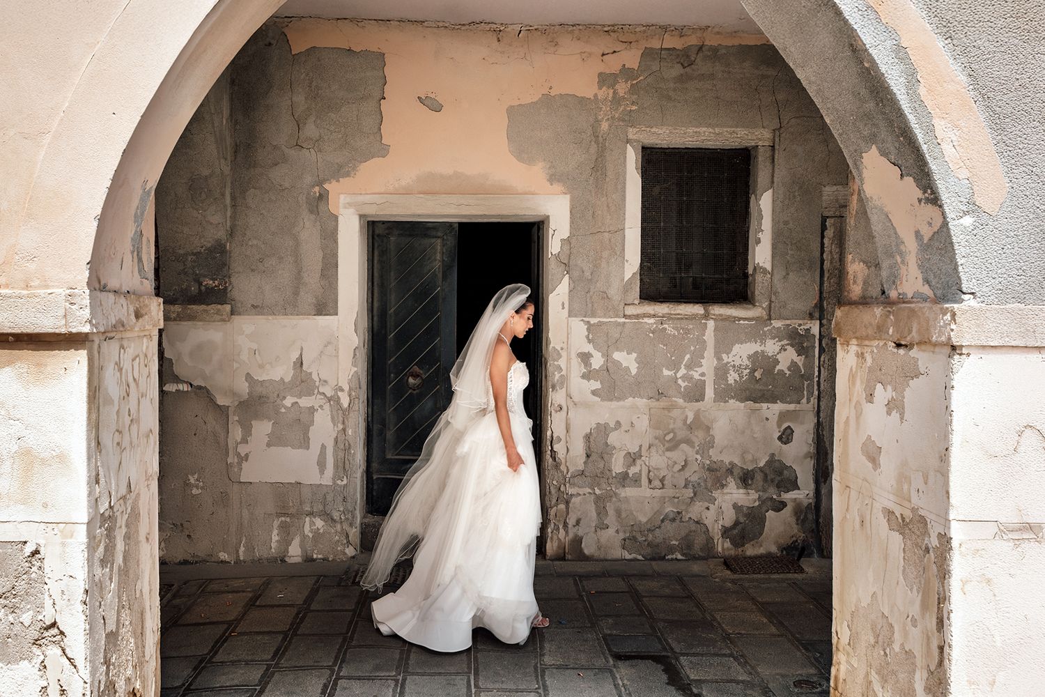 Bride in flowing white gown poses under stone archway in artistic wedding photo.