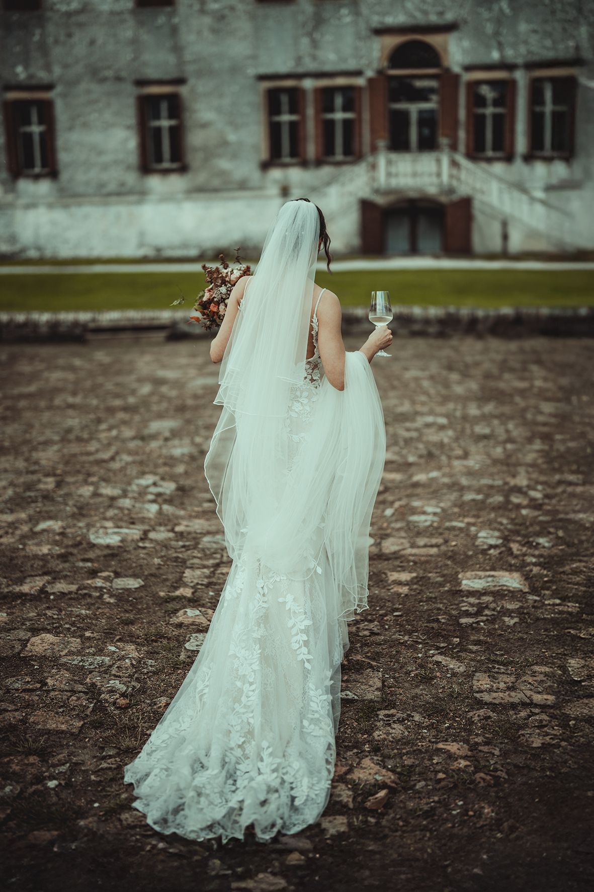 Wedding dress train with intricate lace details photographed against historic stone courtyard.