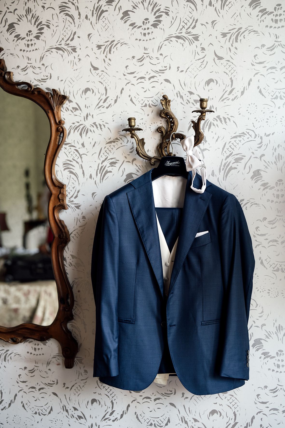Two navy blue suit jackets hanging on ornate hooks against floral wallpaper.