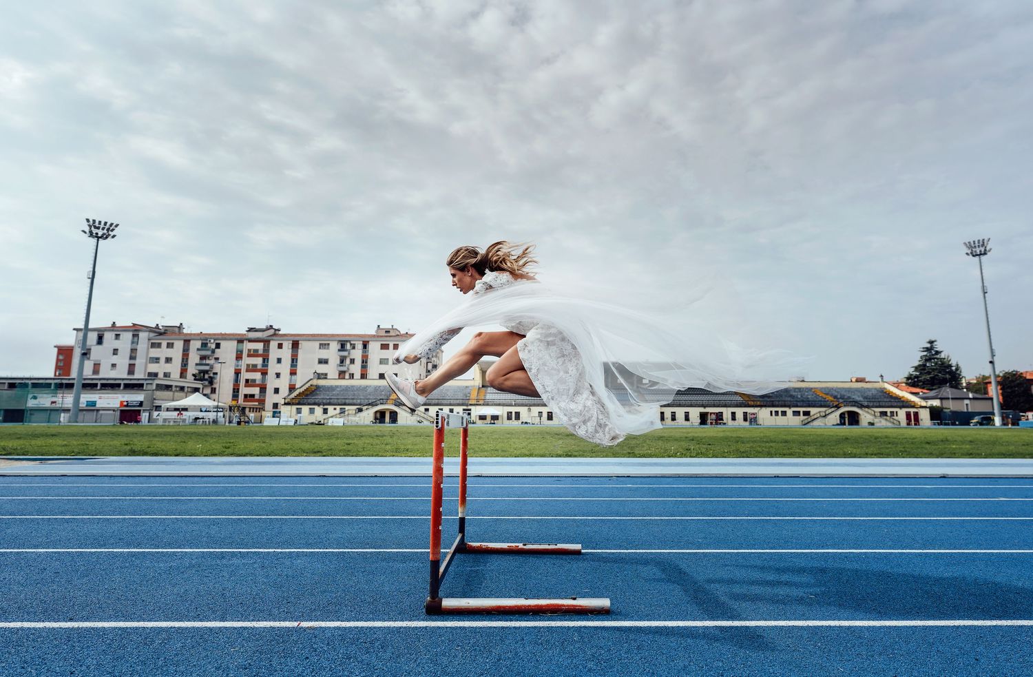 Woman in white dress gracefully leaping over a hurdle on a blue athletic track under a cloudy sky.