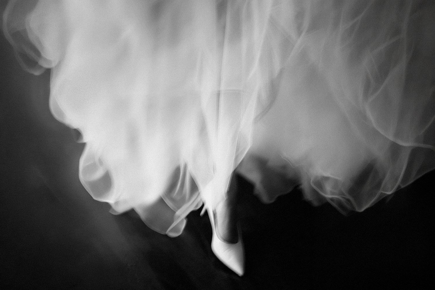 Black and white photo of a bride's white tulle wedding dress hem and heels on a dark floor.