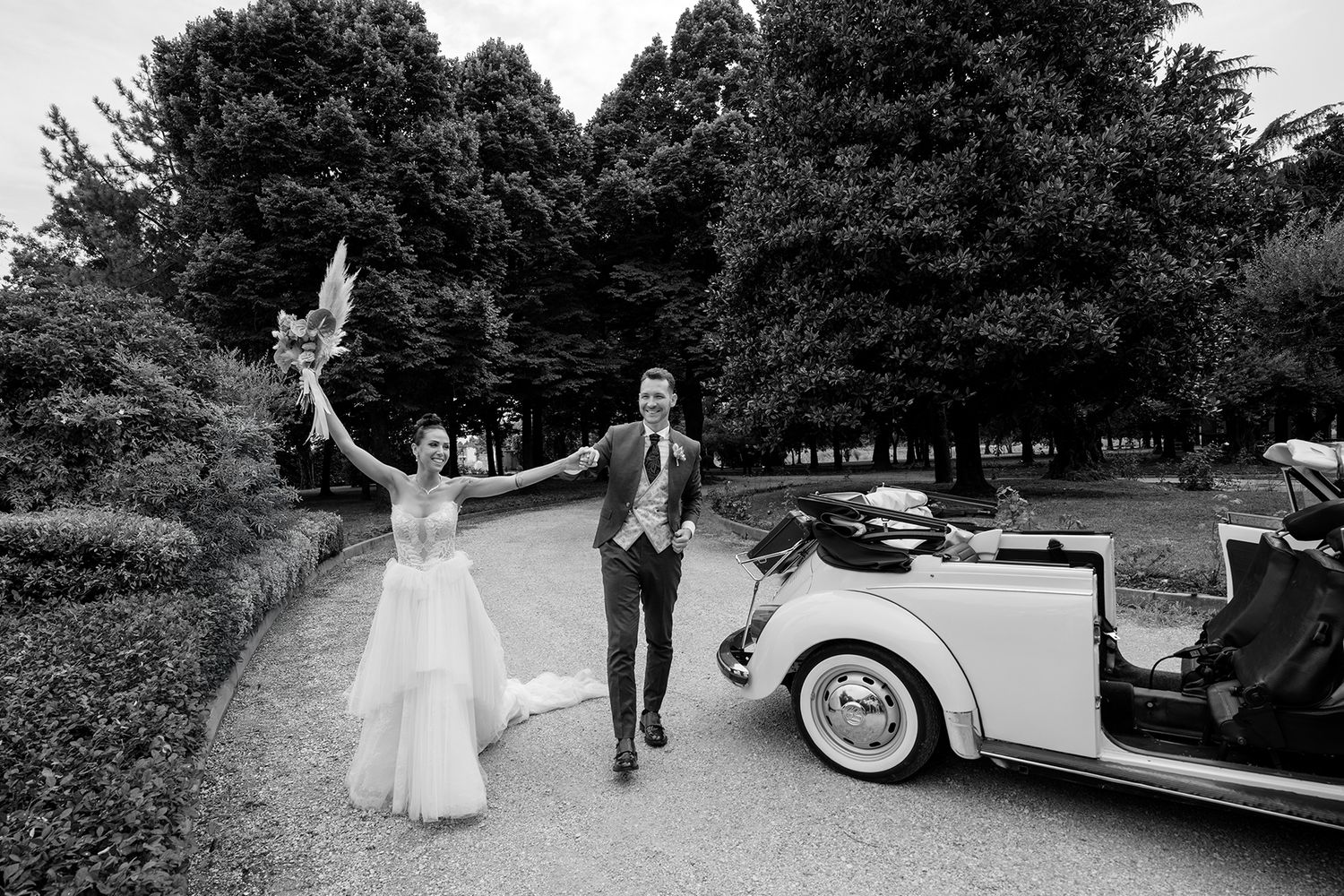 Wedding couple celebrates next to vintage white car on tree-lined path in black and white photo.