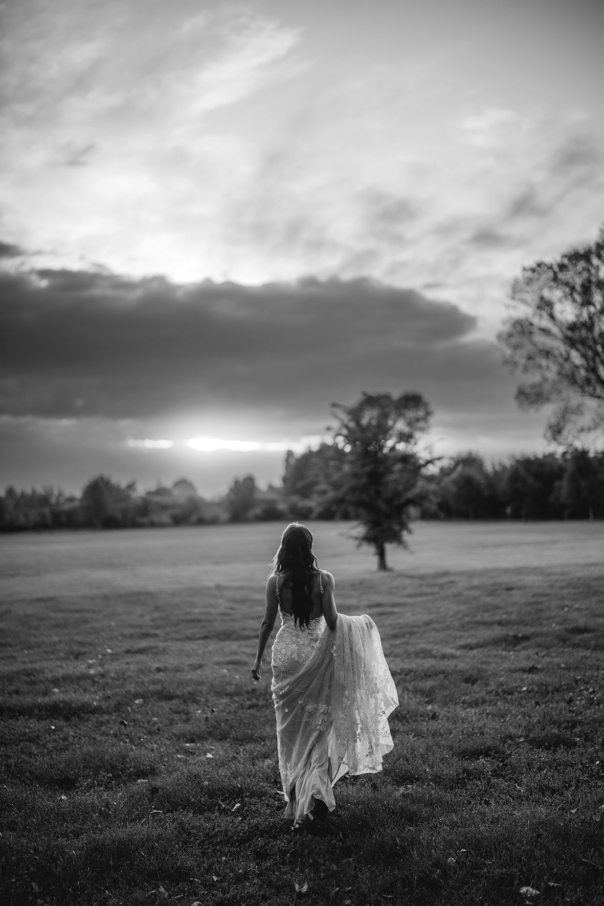 Black and white landscape photo of a figure in flowing dress walking through a misty field at dusk.