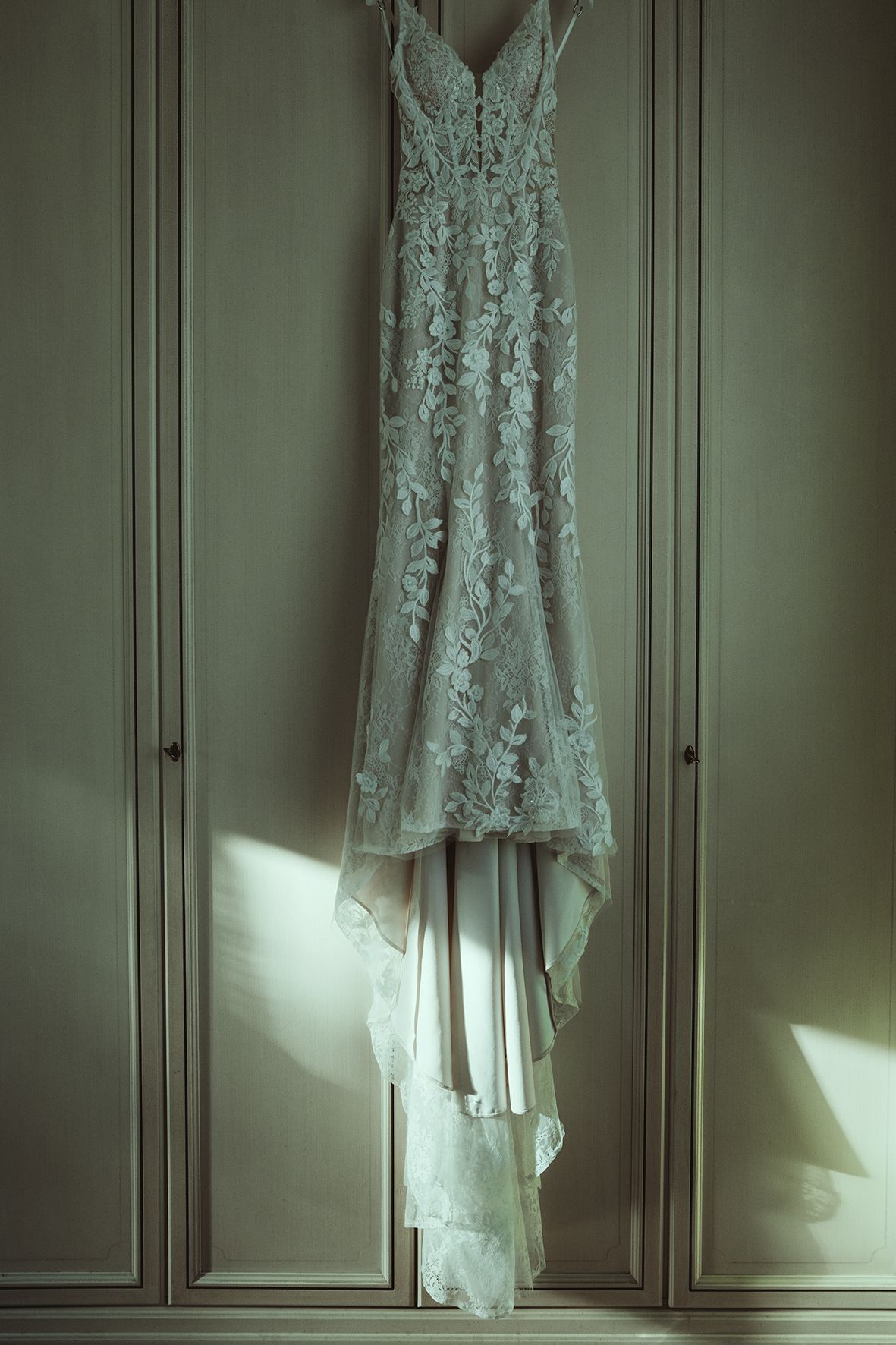 A delicate white lace wedding dress hangs against pale green closet doors.