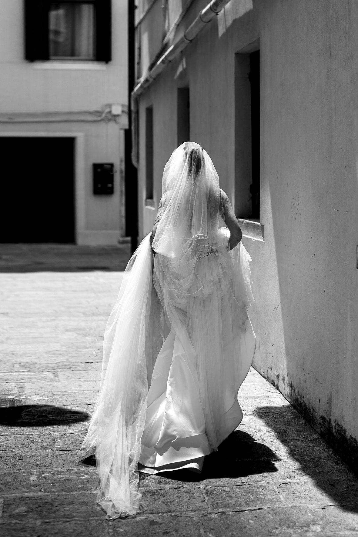 A bride in flowing white dress and veil walks through a narrow alleyway in dramatic black and white.
