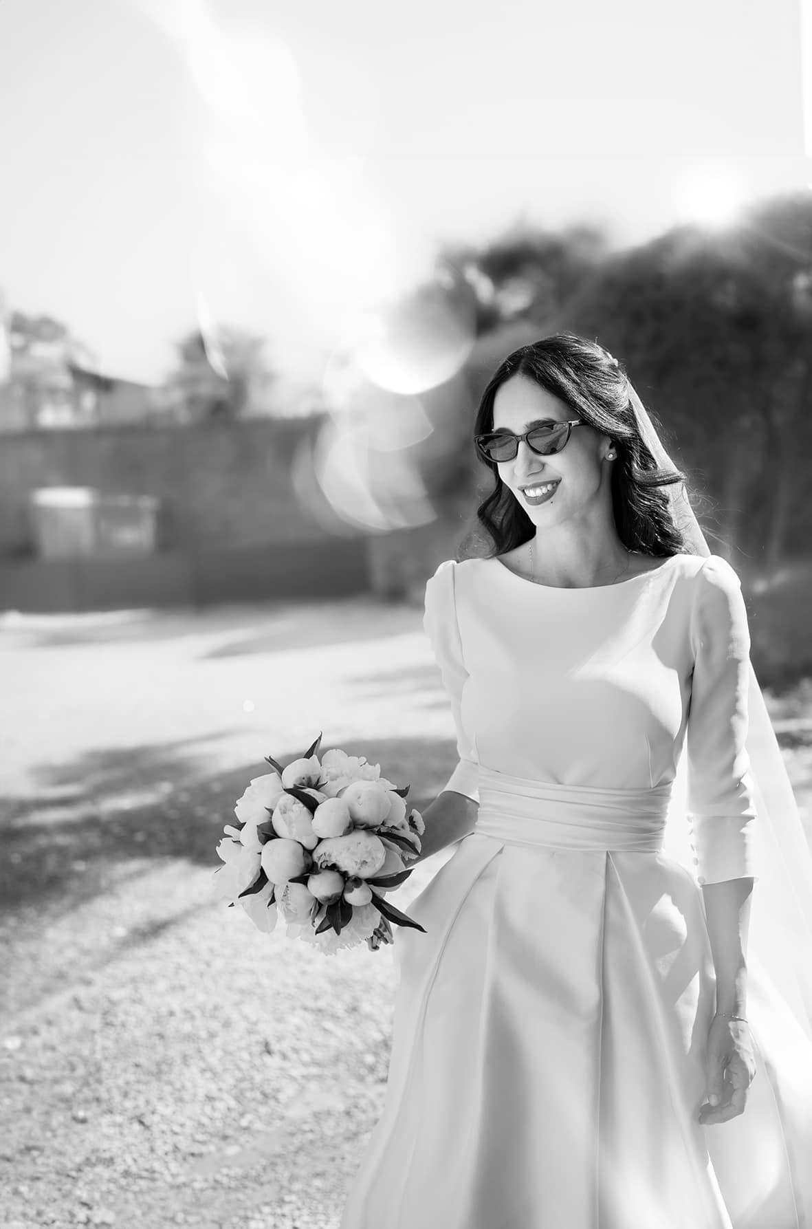 Black and white photo of bride in elegant long sleeve wedding dress and glasses holding bouquet outdoors on sunny day.