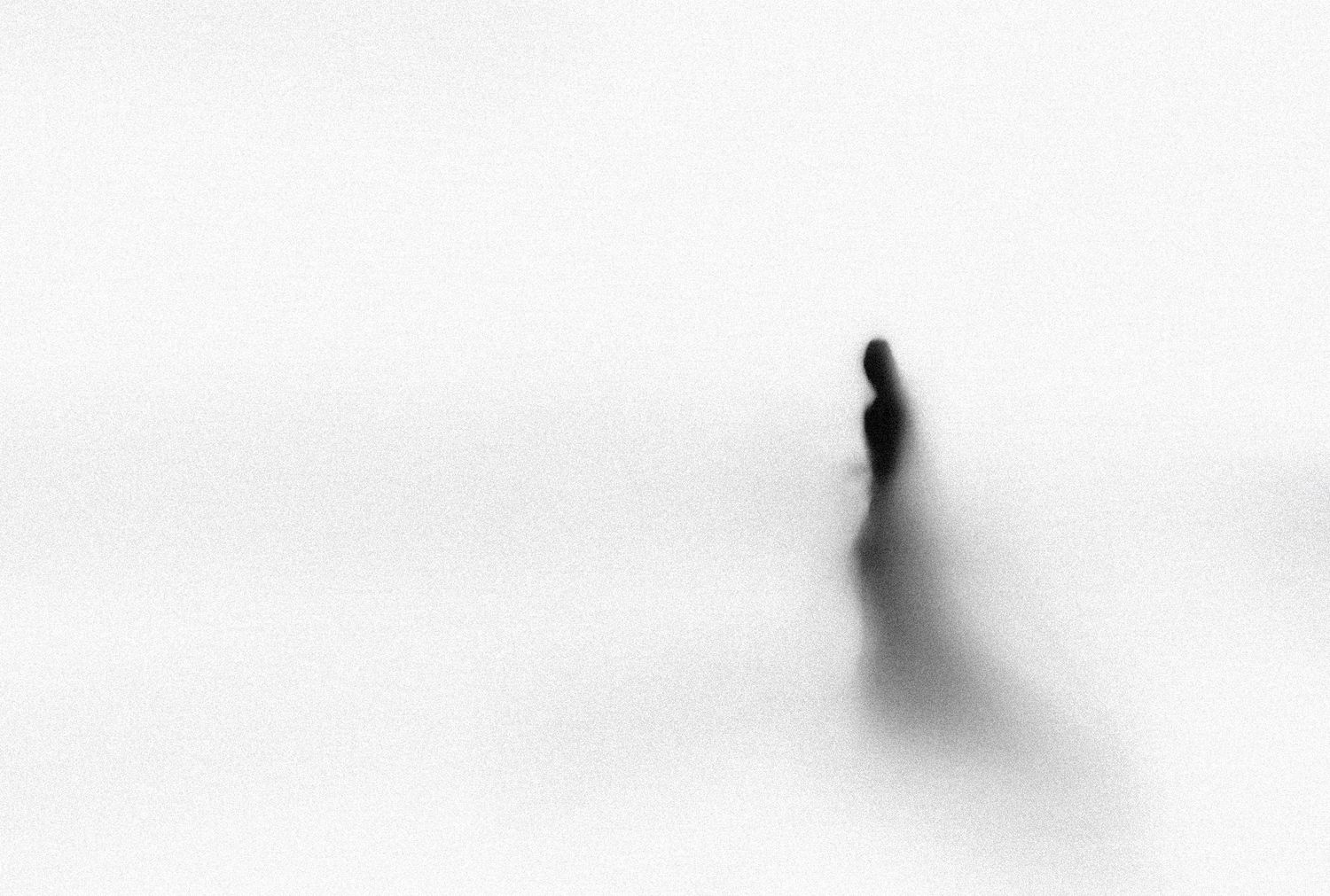 A solitary blurred figure stands in a vast white minimalist landscape in this ethereal black and white photograph.