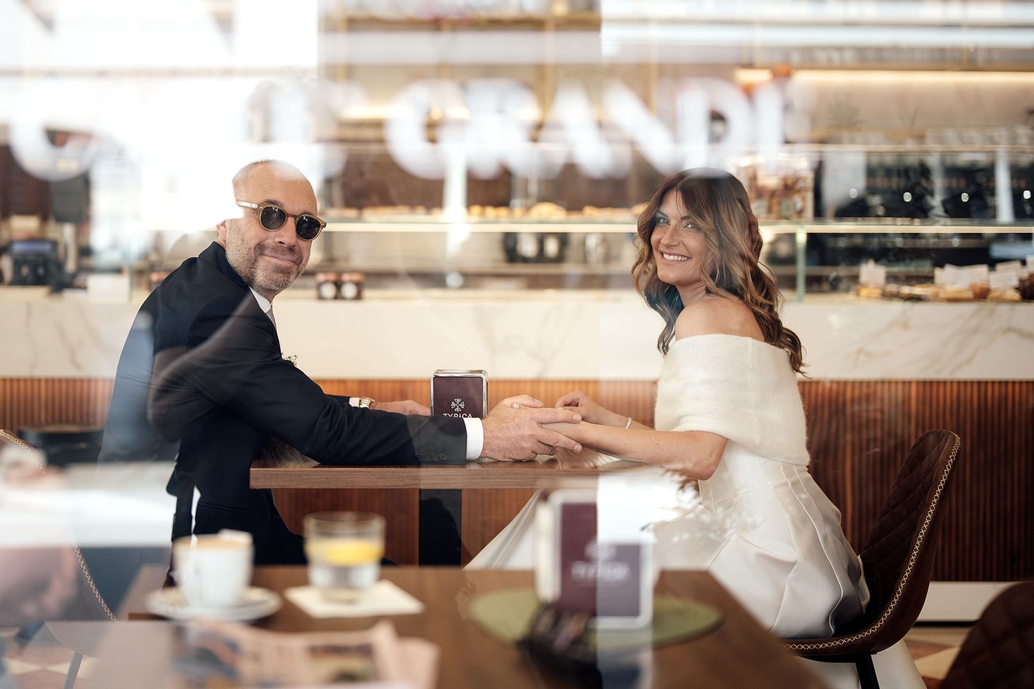 A couple in wedding attire sits at a cafe table sharing an intimate moment.