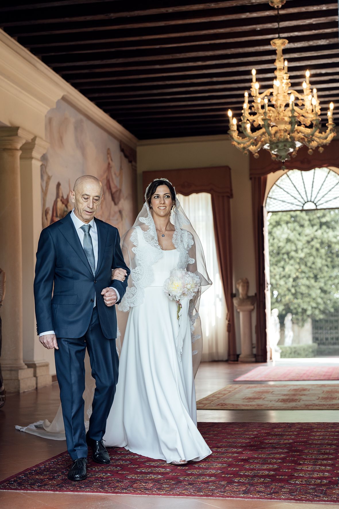 A wedding couple walking through an elegant hallway with chandeliers and a red carpet.