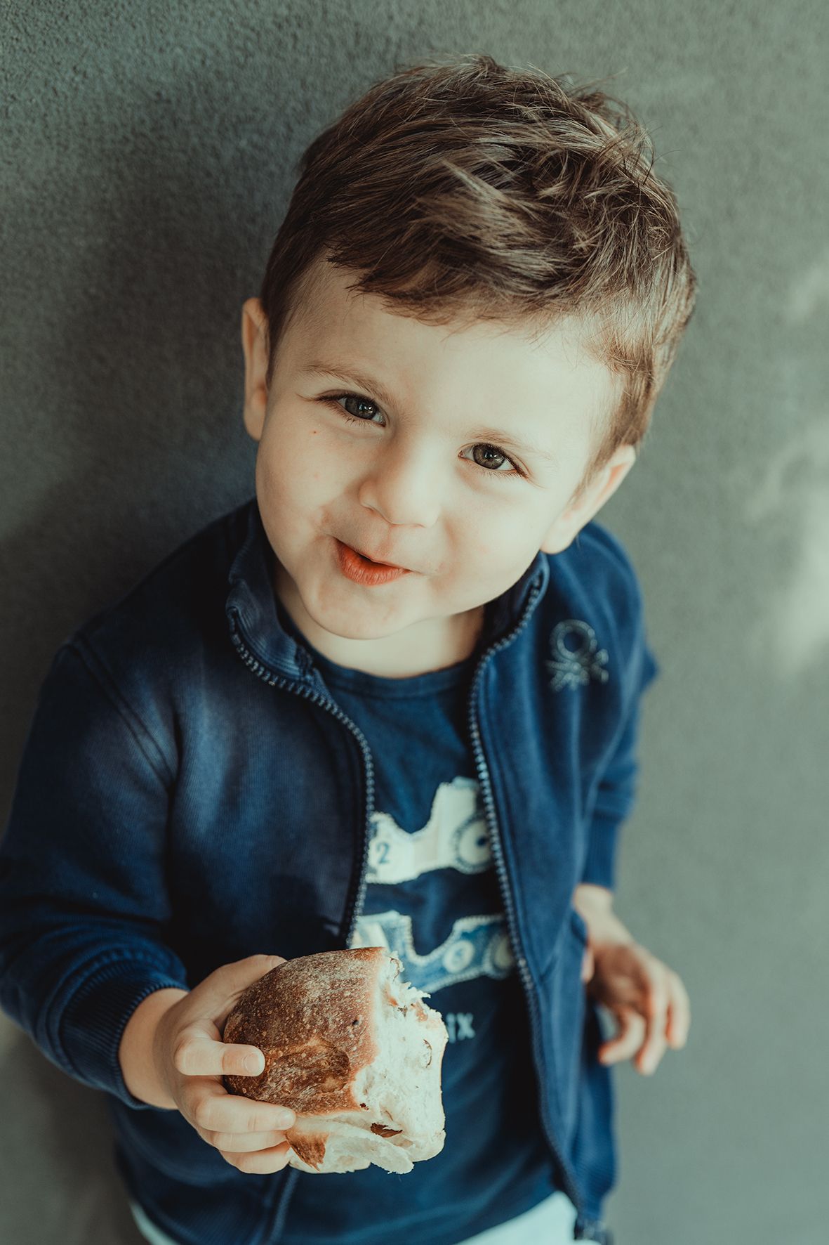Small child in blue jacket enjoying a snack while looking up with a smile.